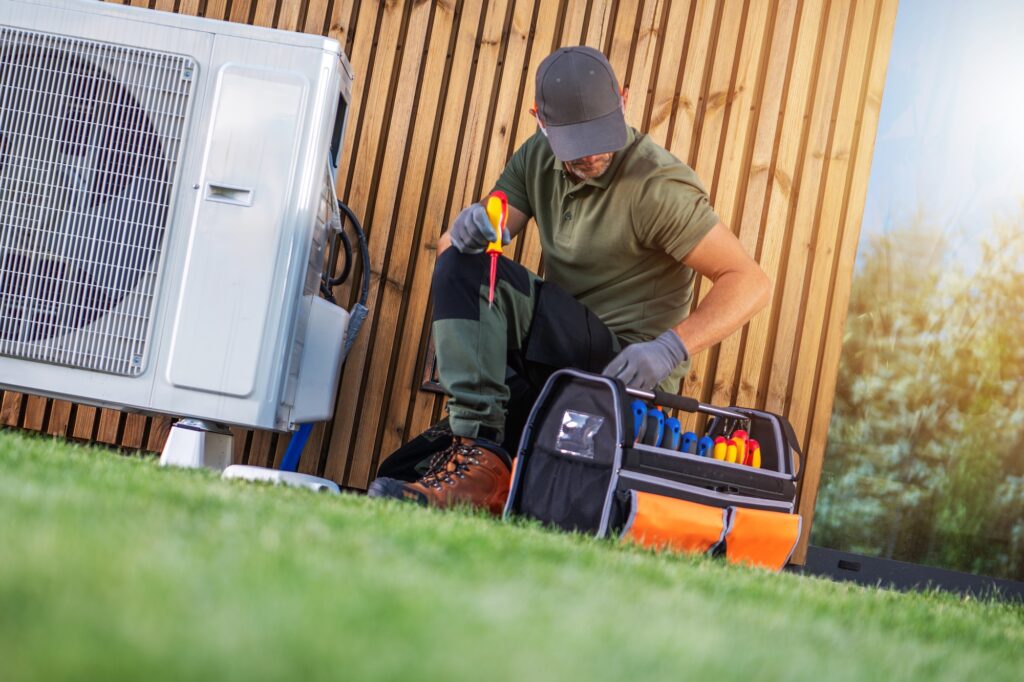 A worker is using tools to repair an air conditioning unit in an outdoor space near a wooden wall and grass.